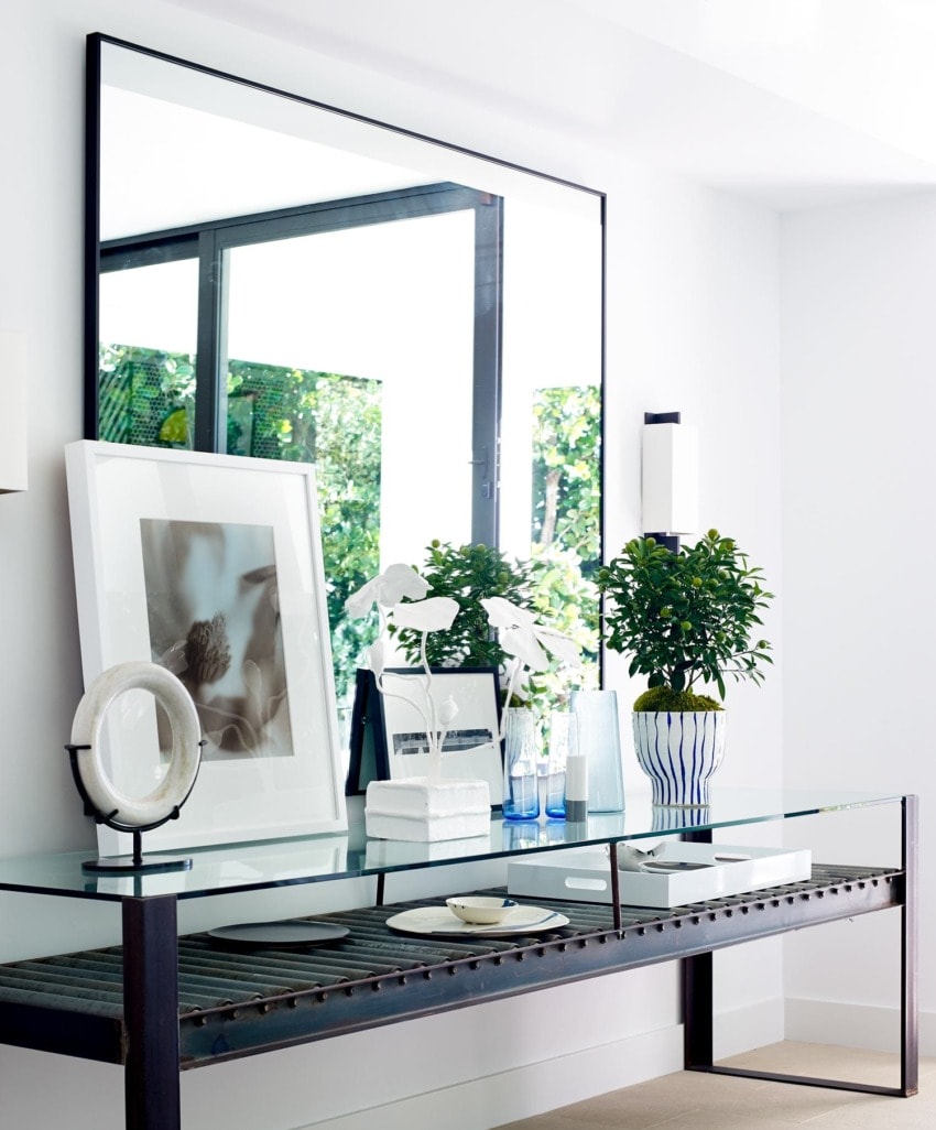 Modern living room with a glass table, decorative items, large framed mirror, and potted plants near a window.