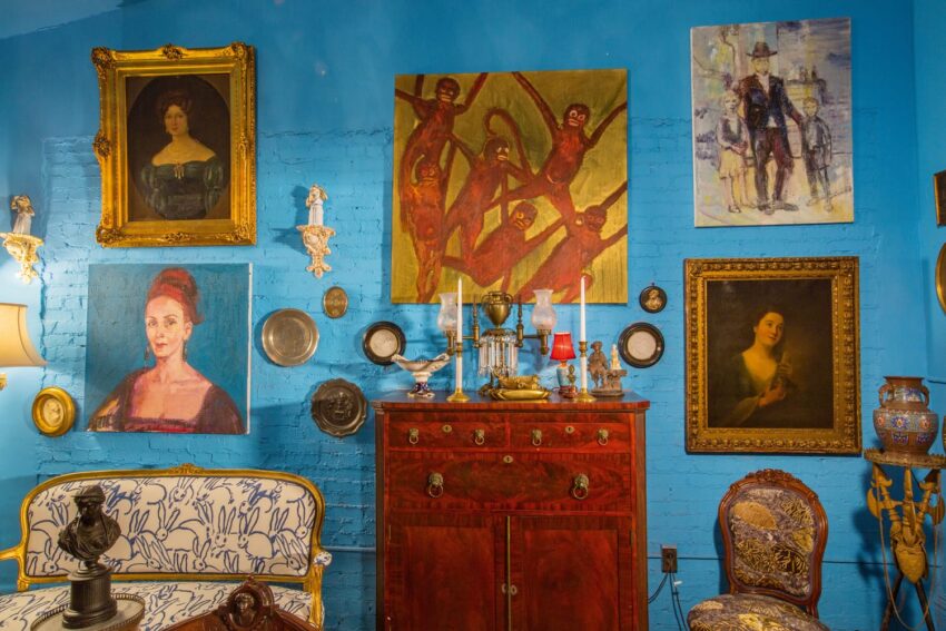 Vintage room with blue walls, eclectic paintings, antique furniture, decorative plates, and ornate chairs.