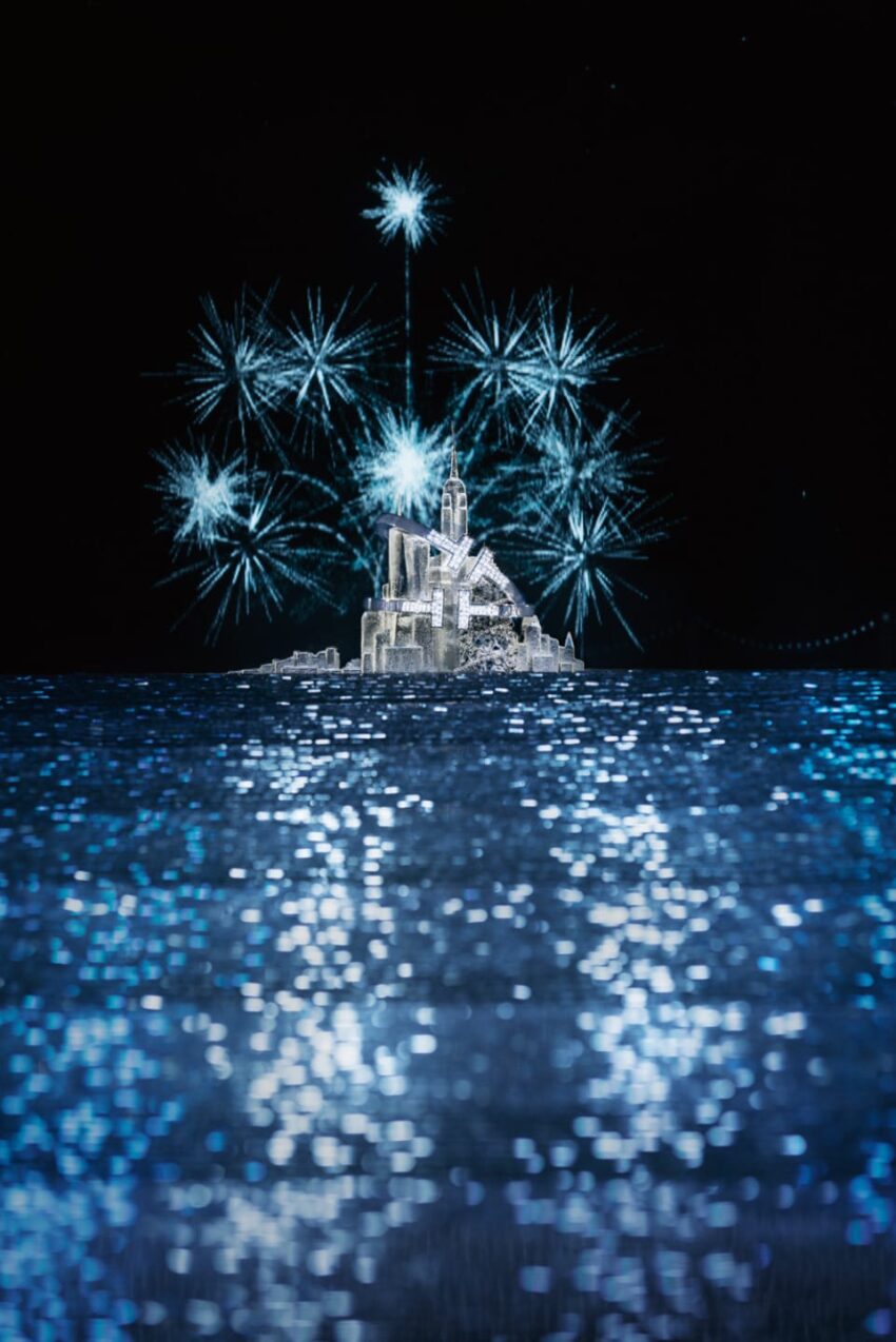 Fireworks illuminate a castle-like structure on a small island, reflecting on a shimmering body of water at night.