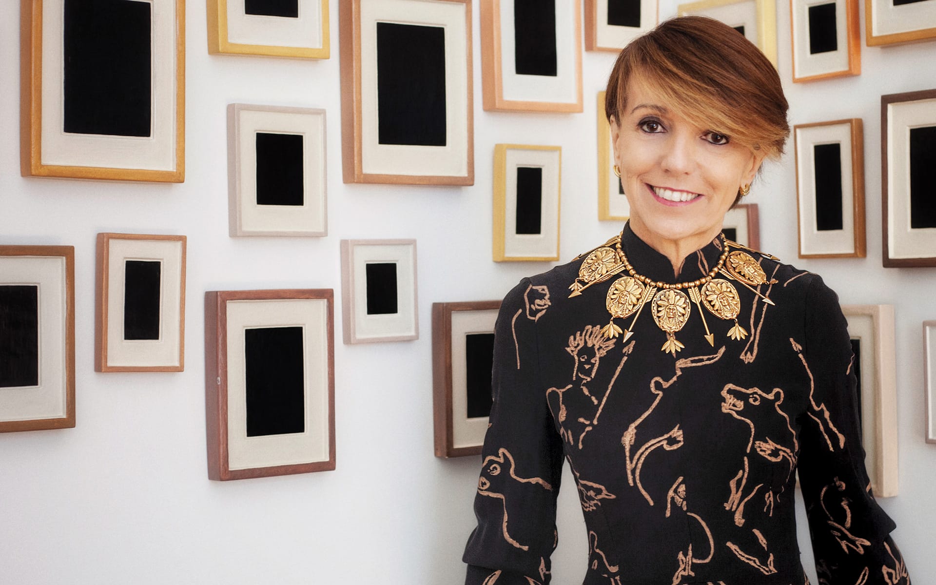 Person in black designer outfit with gold accents, smiling in front of a wall adorned with various empty picture frames.