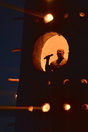 Person singing on an illuminated stage inside a cylindrical structure with circular openings at night.