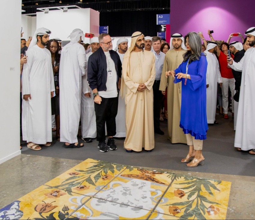 Group of people in traditional and modern attire viewing artwork on the floor at an exhibition.