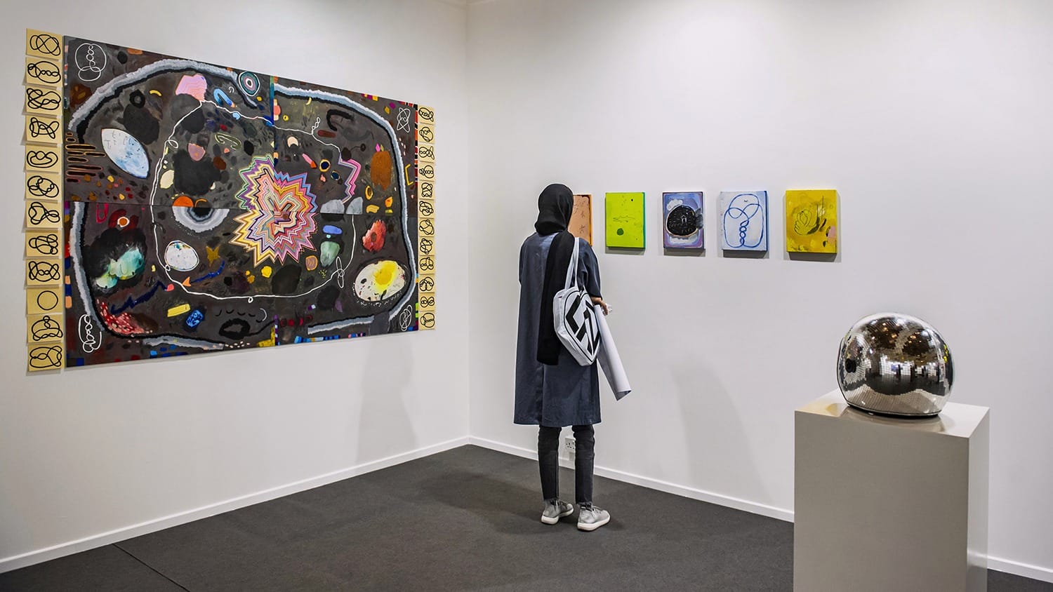 Person observing colorful abstract paintings in a gallery with a reflective sphere on a pedestal nearby.