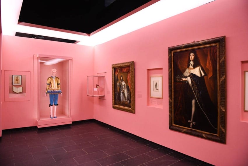 Museum gallery with vibrant pink walls displaying historical portraits and a mannequin in colorful attire.