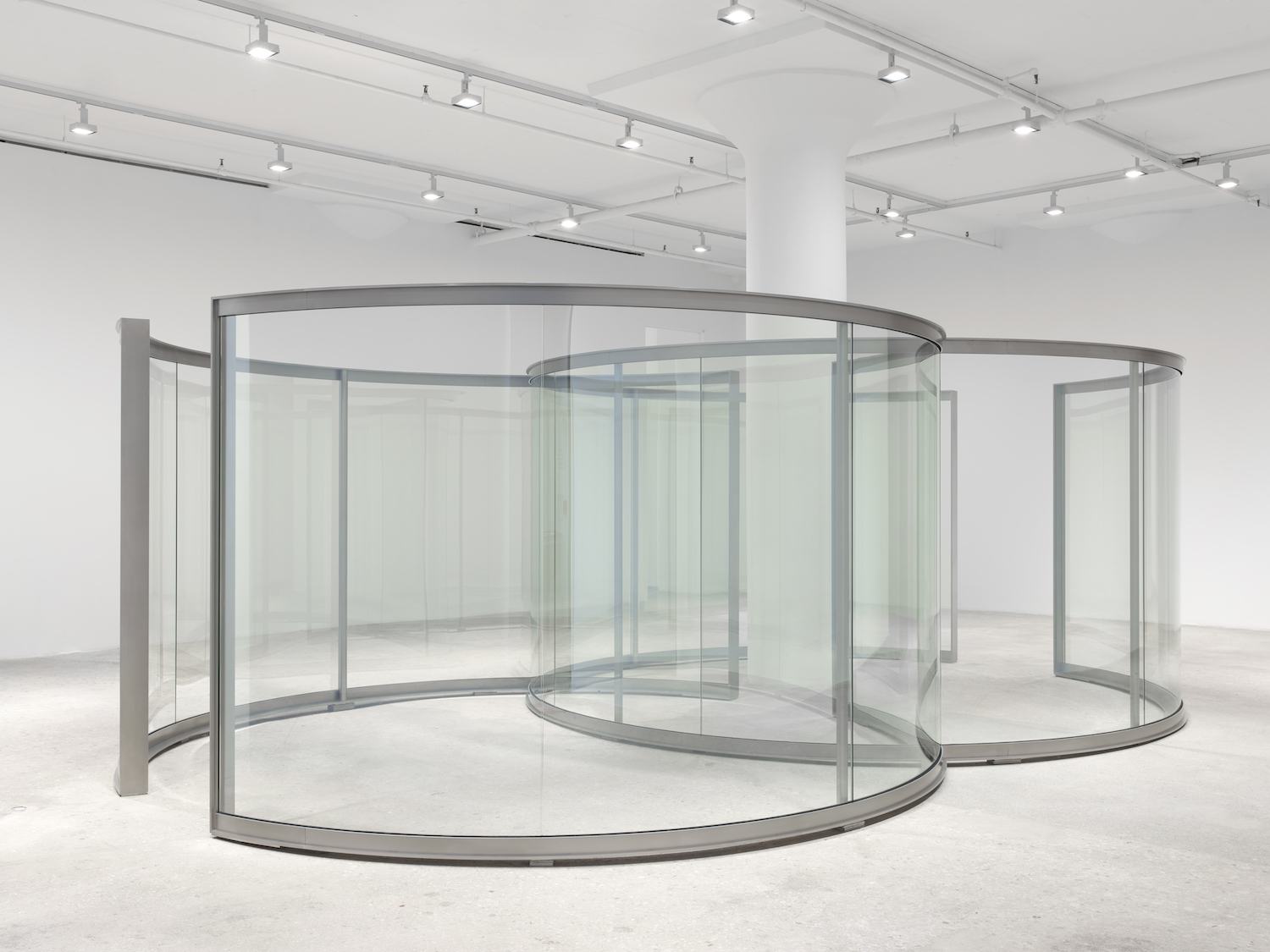 Modern art installation featuring curved glass panels and gray frames in a white-walled gallery space with overhead lighting.