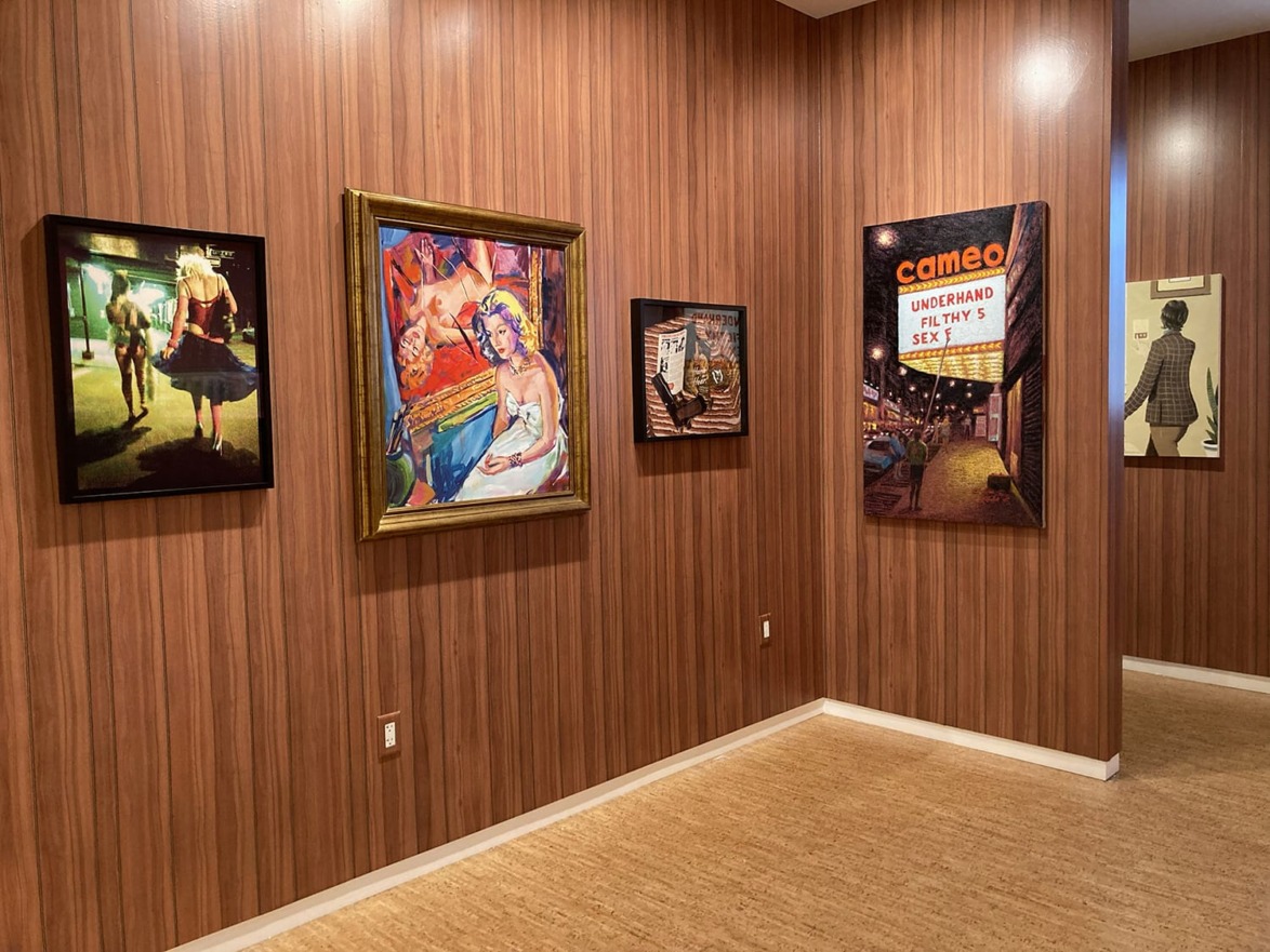 Art gallery with various framed paintings on wooden walls, including colorful portraits and a movie poster.