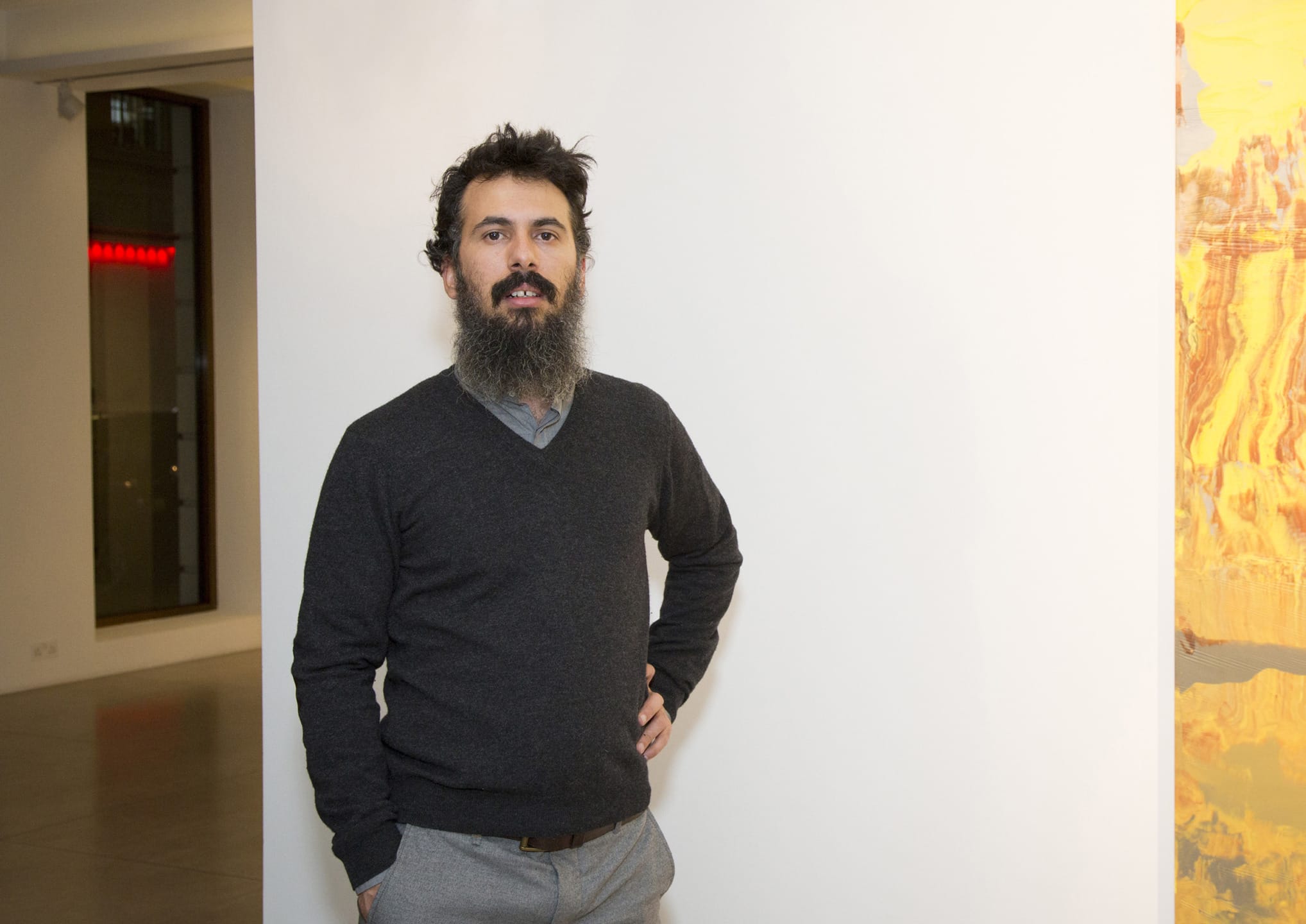 Man with a beard standing in a modern art gallery, wearing a dark sweater and gray pants, with abstract art visible in the background.