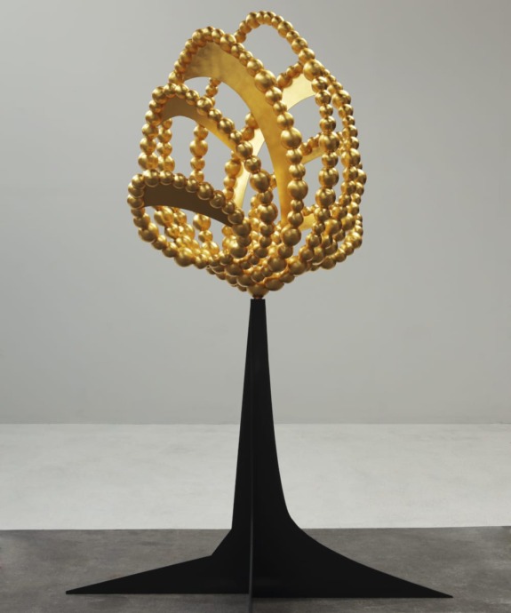 Modern gold spherical sculpture on slender black stand, displayed in a minimalistic gallery space.