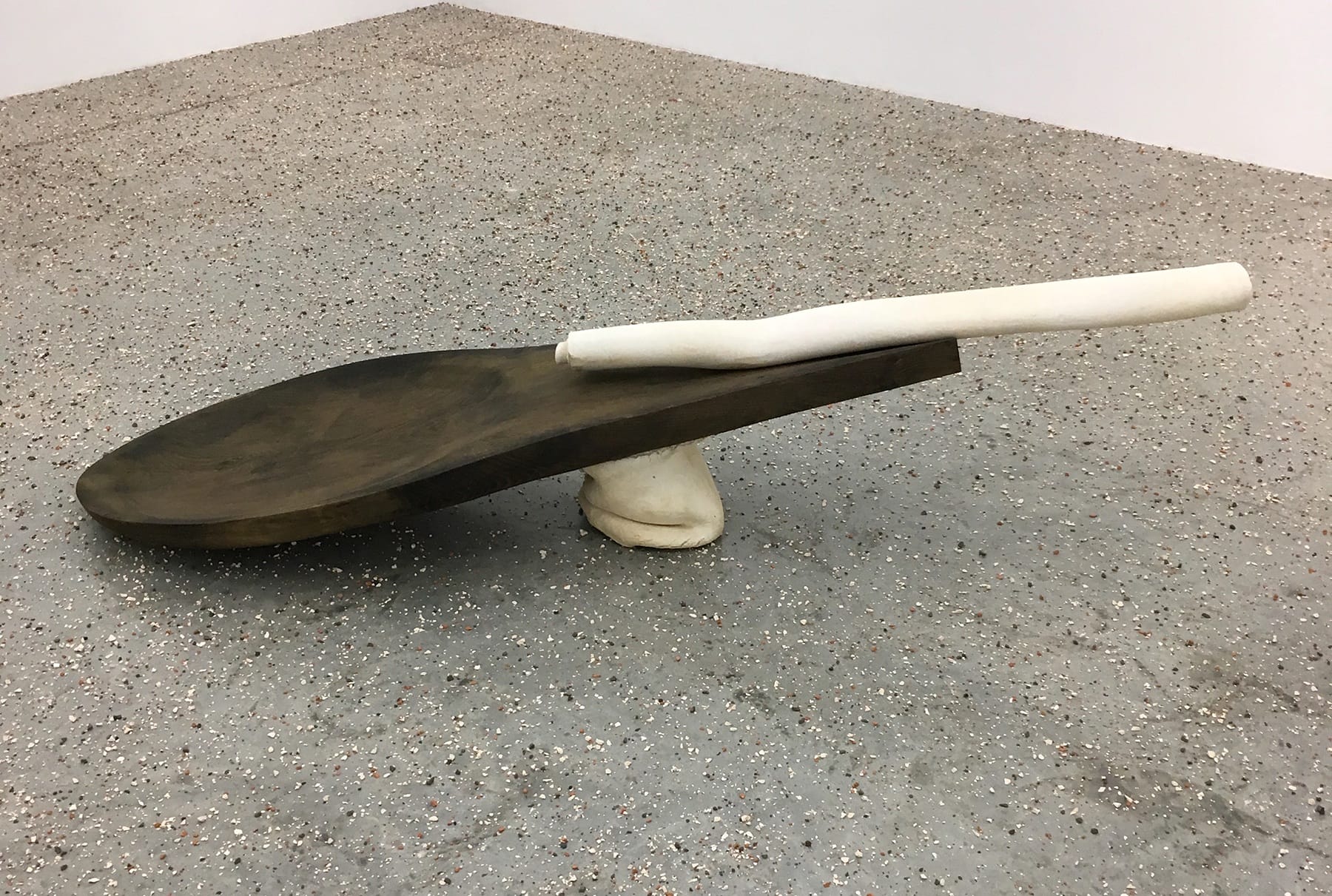 Large abstract wooden sculpture resembling a spoon, with a textured handle resting on a concrete floor in a gallery setting.