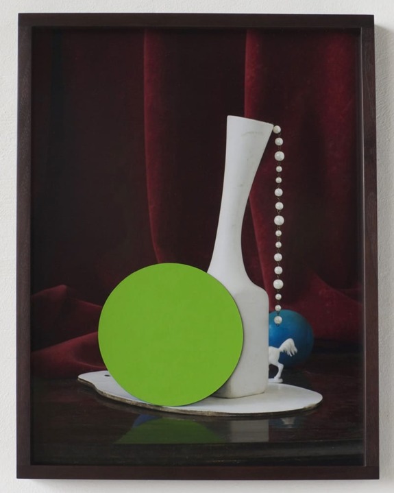 Decorative still life with a tall white vase, green circle, pearls, and a small horse figurine against a red velvet backdrop.