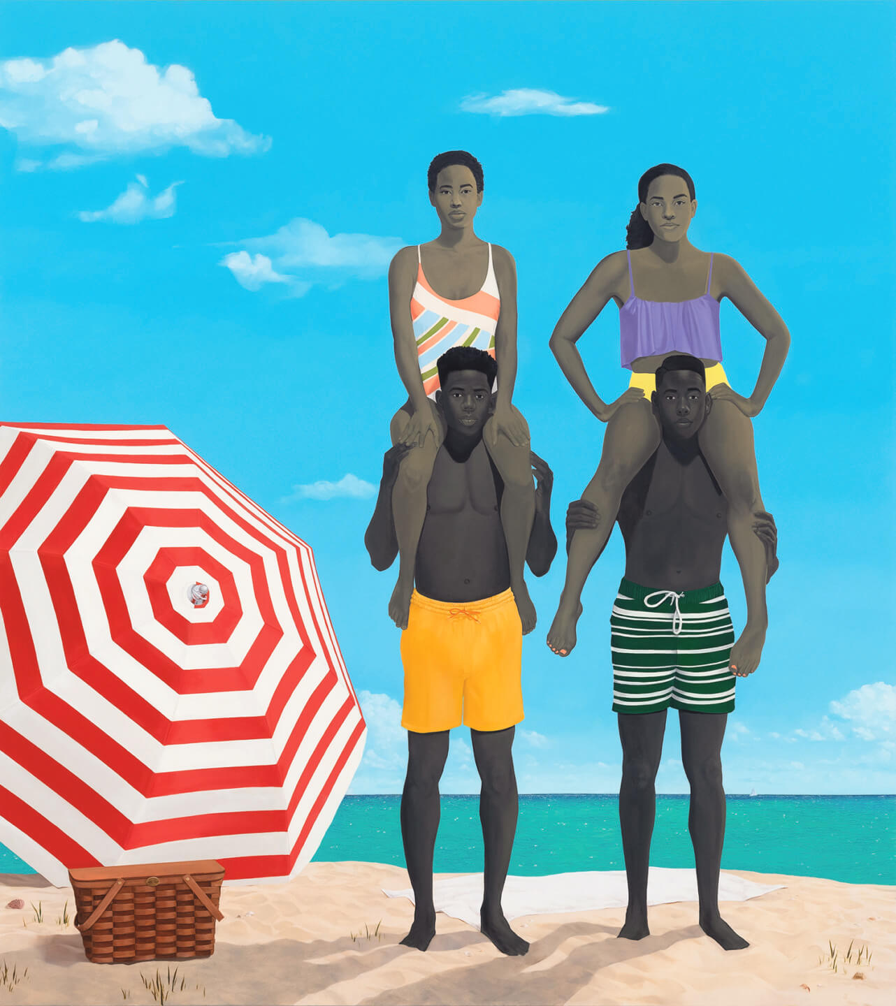 Two people standing on a beach holding others on their shoulders, next to a red and white striped umbrella and a picnic basket.