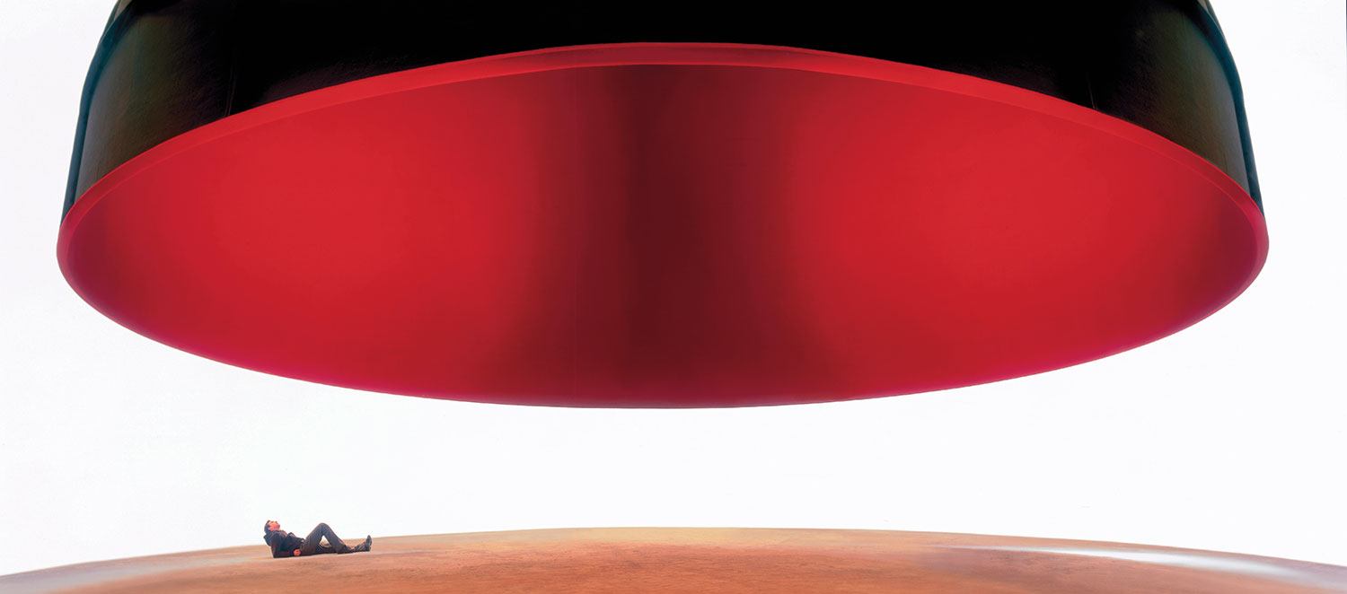 Person lying under a large, suspended circular object with a black exterior and red underside in a minimalist setting.