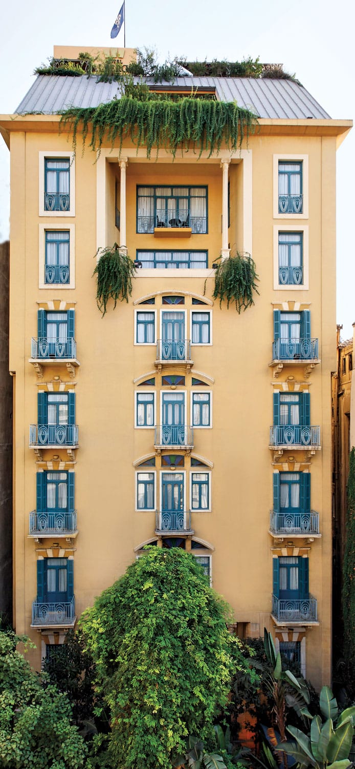 Yellow building with blue window shutters, green plants cascading from balconies, and trees at the base.