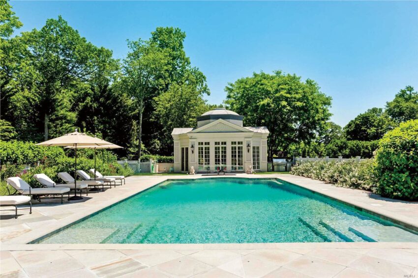 Luxurious outdoor swimming pool with sun loungers, large umbrellas, surrounded by lush greenery and elegant pool house.