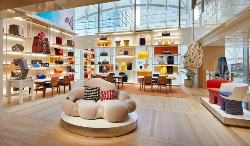 Luxury retail store interior with colorful handbags on shelves, modern furniture, and large windows letting in natural light.