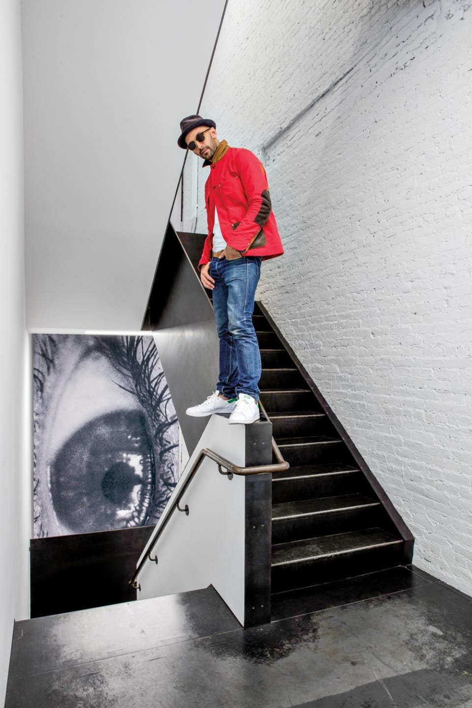 Man wearing a red jacket and hat standing on a staircase with eye mural on the wall behind him.