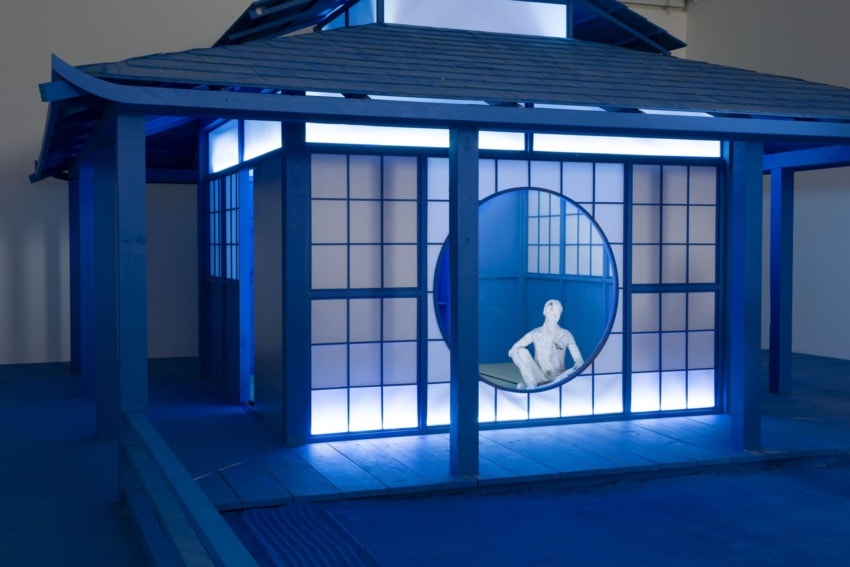 A blue-lit structure with shoji-style windows and a circular opening with a seated statue inside.