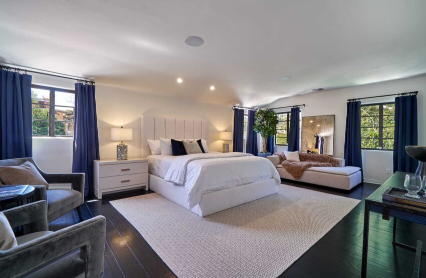 Spacious modern bedroom with a king-sized bed, cozy seating area, dark wood flooring, and large windows with blue curtains.