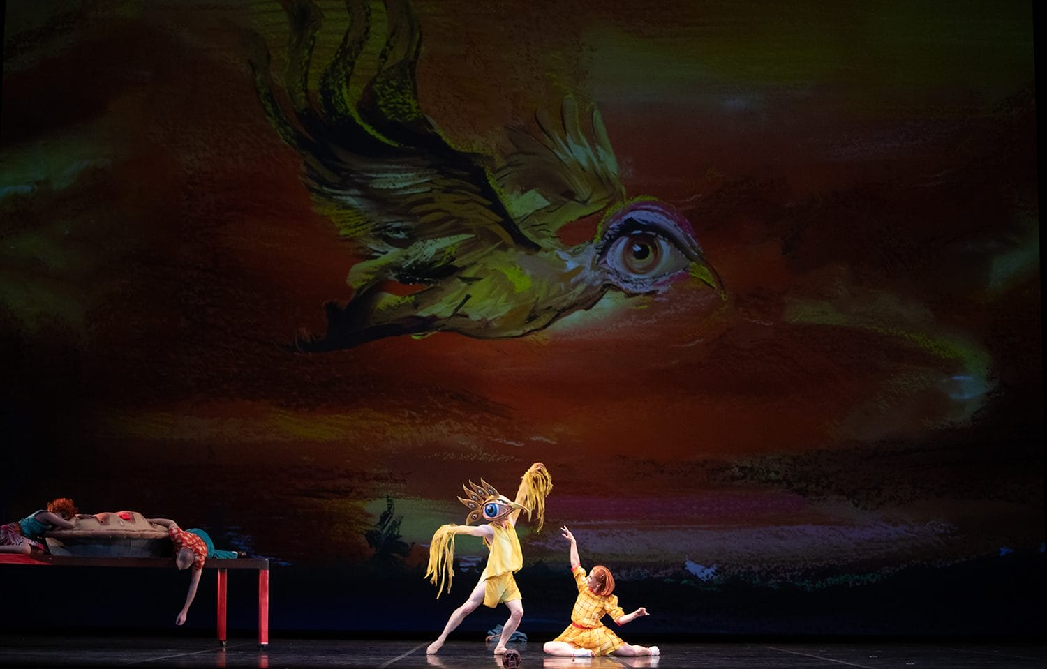 Performers in colorful costumes on stage with a large bird art backdrop in a dramatic scene.