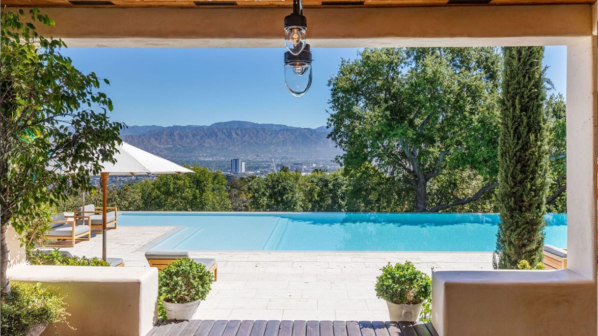 View of infinity pool with city and mountain backdrop, framed by trees and plants, under a wooden pergola.
