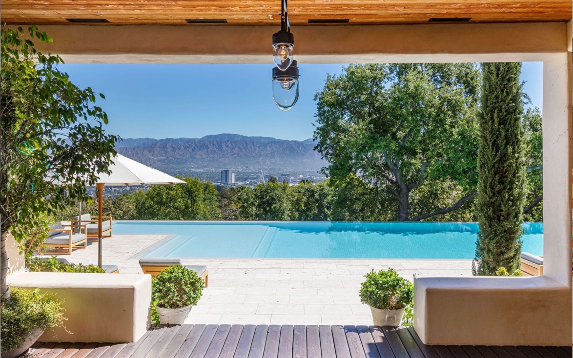 View of infinity pool with city and mountain backdrop, framed by trees and plants, under a wooden pergola.