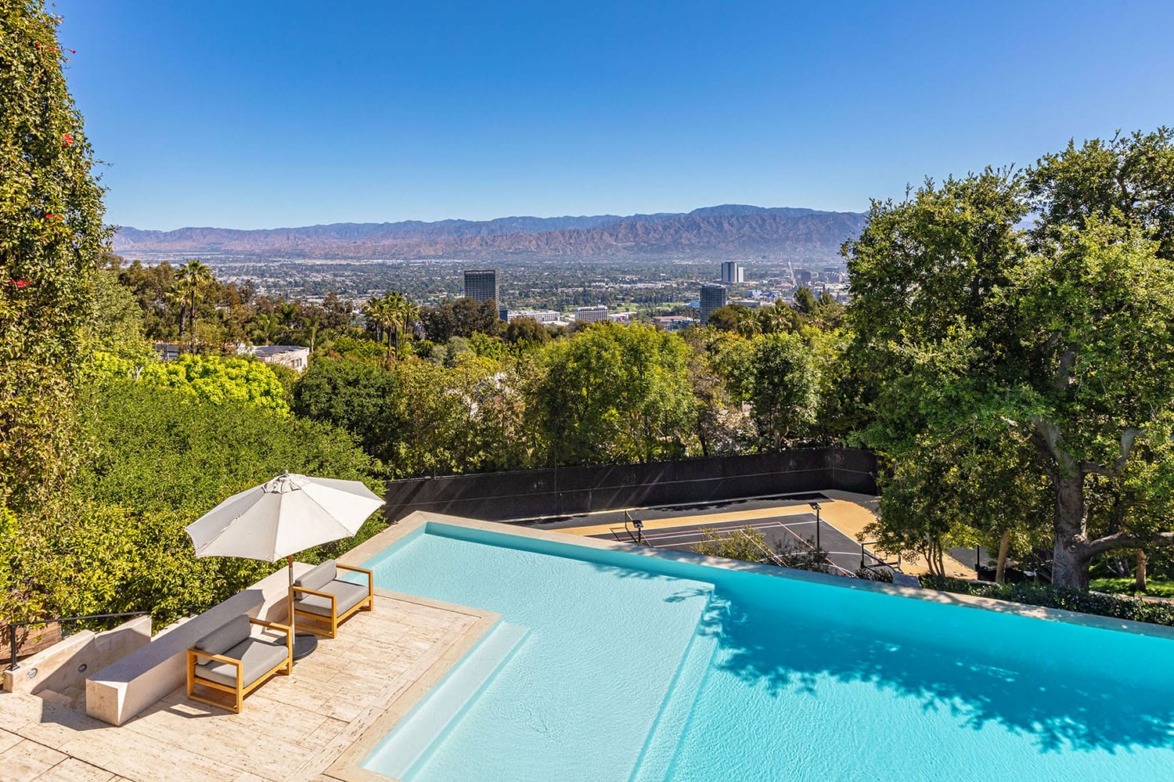 Infinity pool overlooking a cityscape with trees and mountains in the background, two lounge chairs under an umbrella.