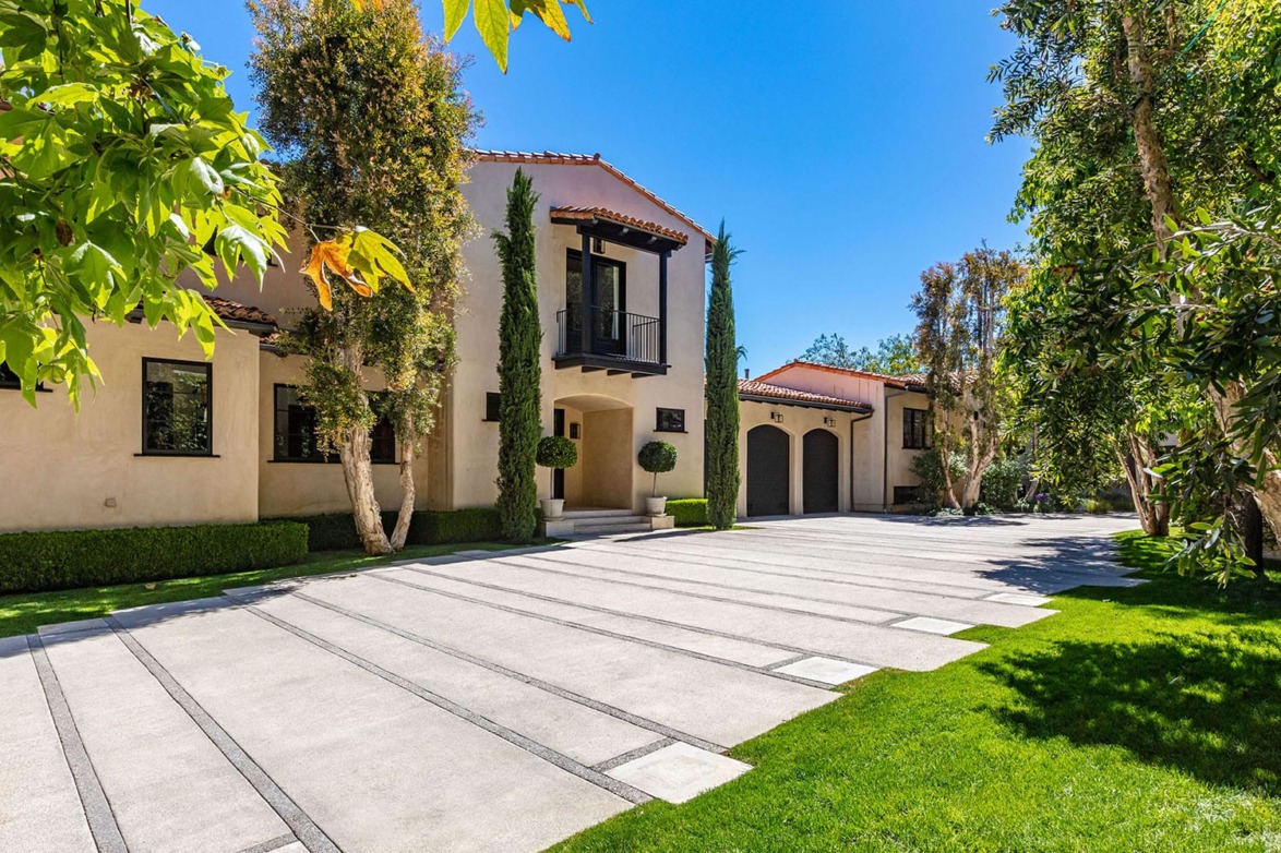Luxury Mediterranean-style house with tall trees, paved driveway, and lush greenery under a bright blue sky.