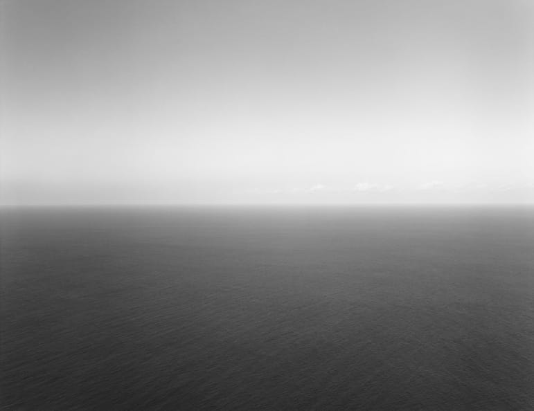 Black and white photo of a calm, expansive ocean meeting a clear sky, showing the serene beauty of a seascape horizon.