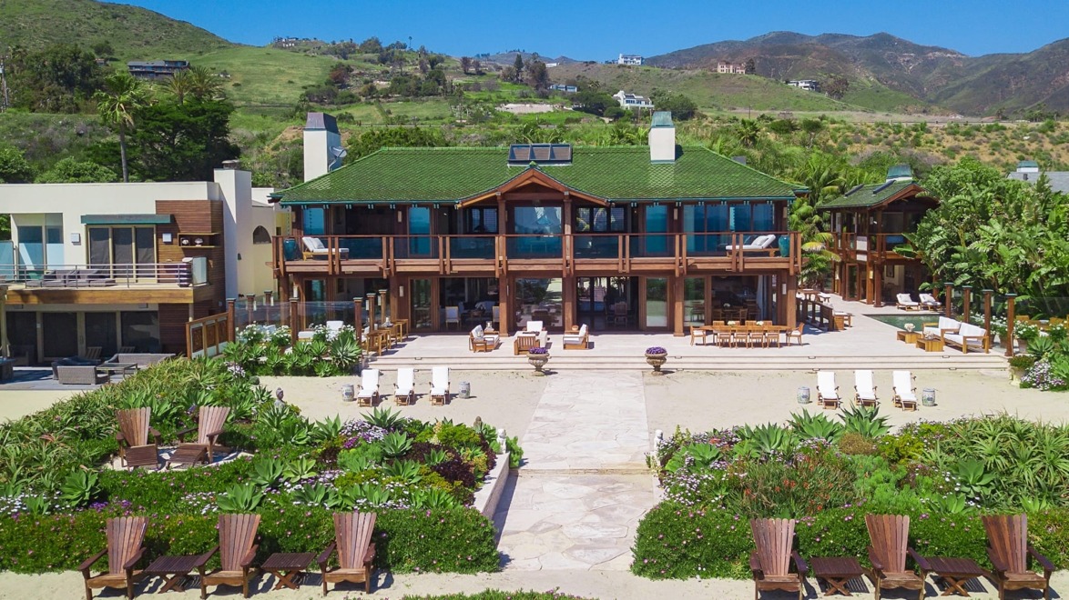 Large beachfront house with green roof, surrounded by lush plants, wooden deck chairs, and scenic hills in the background.