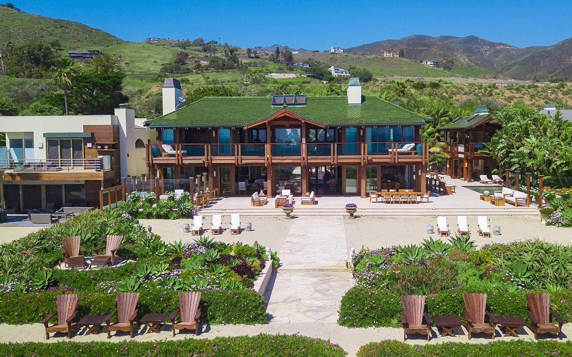 Large beachfront house with green roof, surrounded by lush plants, wooden deck chairs, and scenic hills in the background.