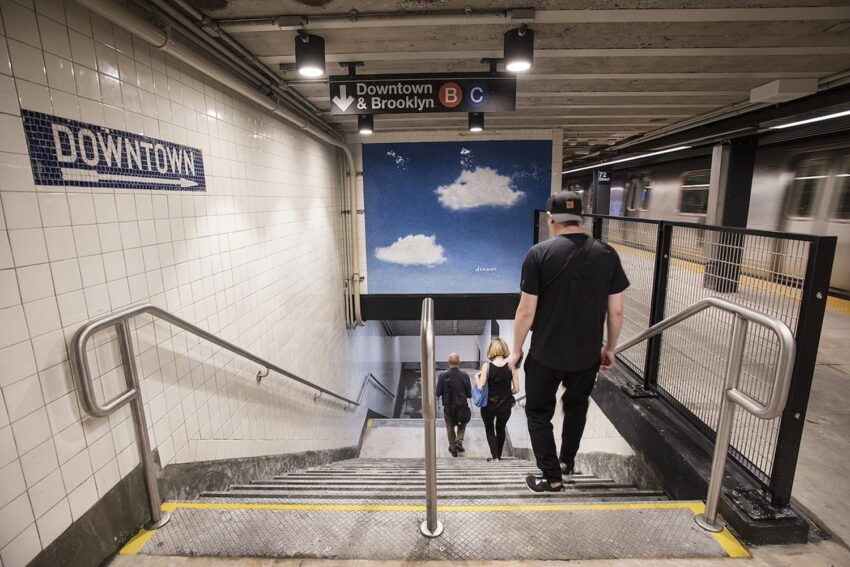 People descending stairs in a subway station with a blue mural featuring clouds and a train arriving on the platform.