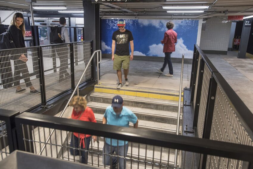 People walking in a subway station, with steps leading down and a mural of a clouded blue sky in the background.