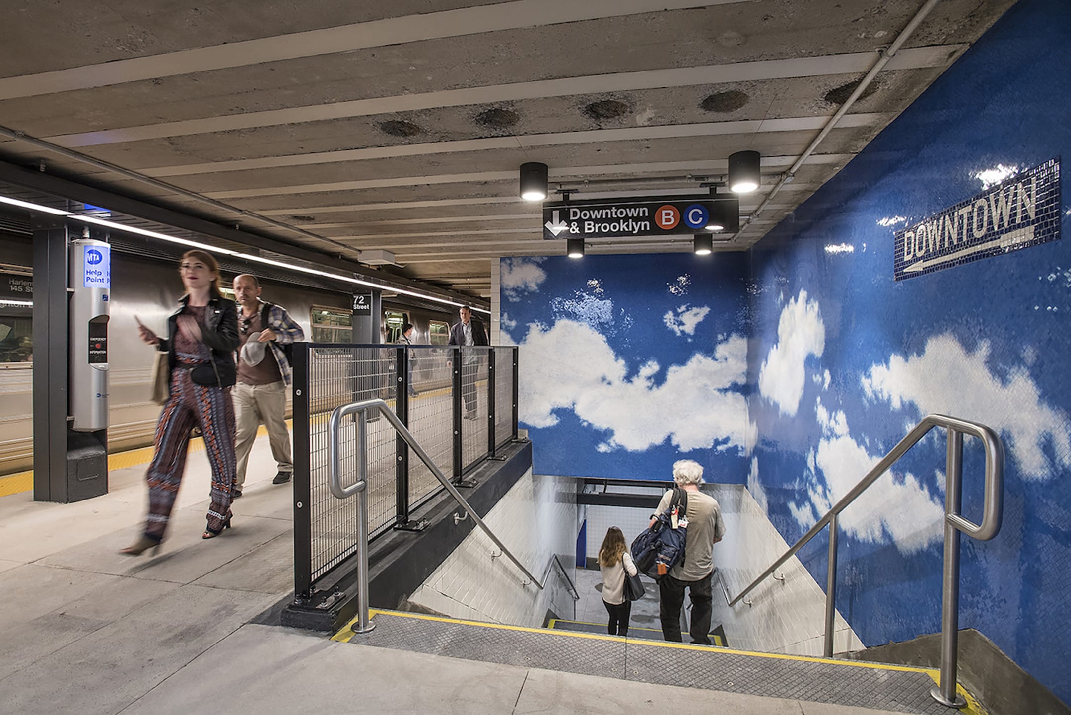 Subway station entrance with stairs, mural of sky on wall, people walking, and signs for Downtown & Brooklyn, B C trains.