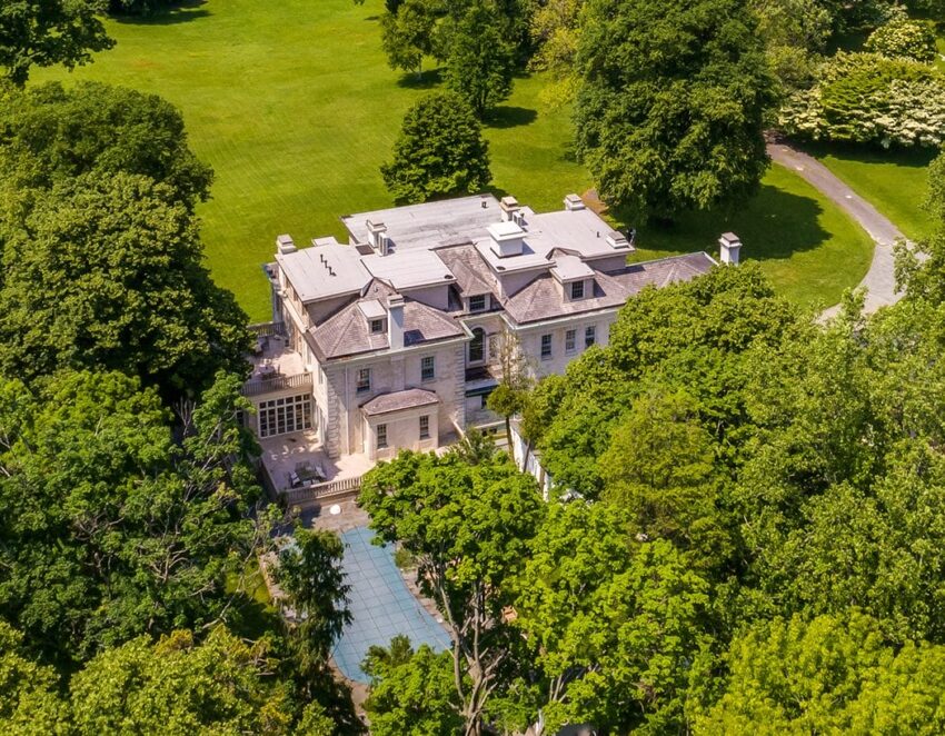 Aerial view of a large historic mansion surrounded by lush greenery and trees with a clear pool in the foreground.