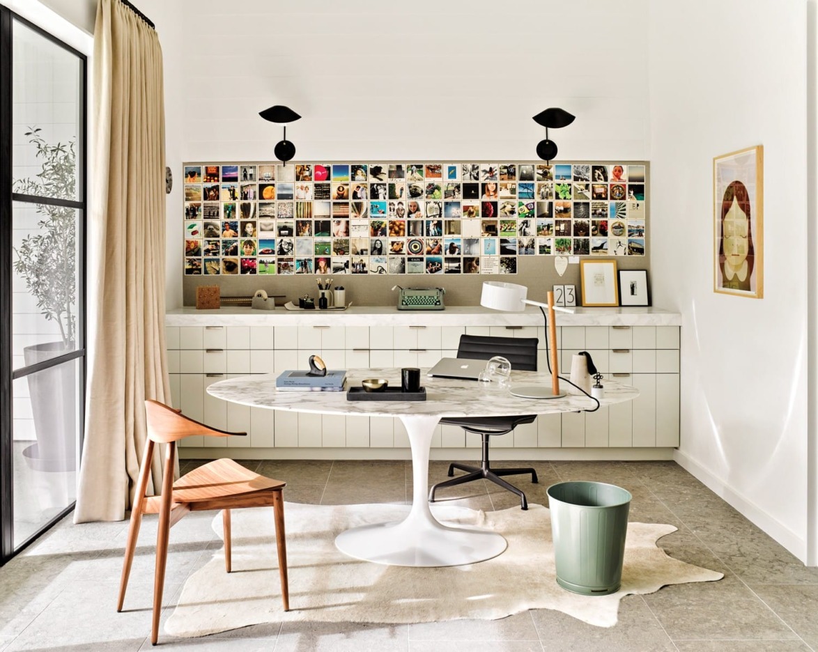 Modern home office with round white table, wooden chair, wall collage of photos, and natural light streaming through glass doors.