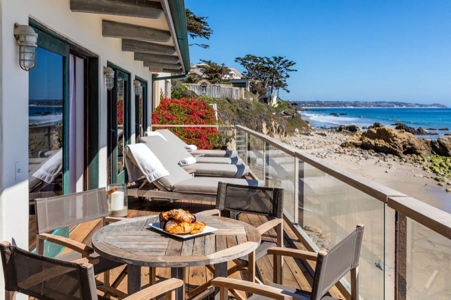 Beachfront patio with wooden furniture, croissants on a table, and lounge chairs overlooking the ocean and rocky shore.