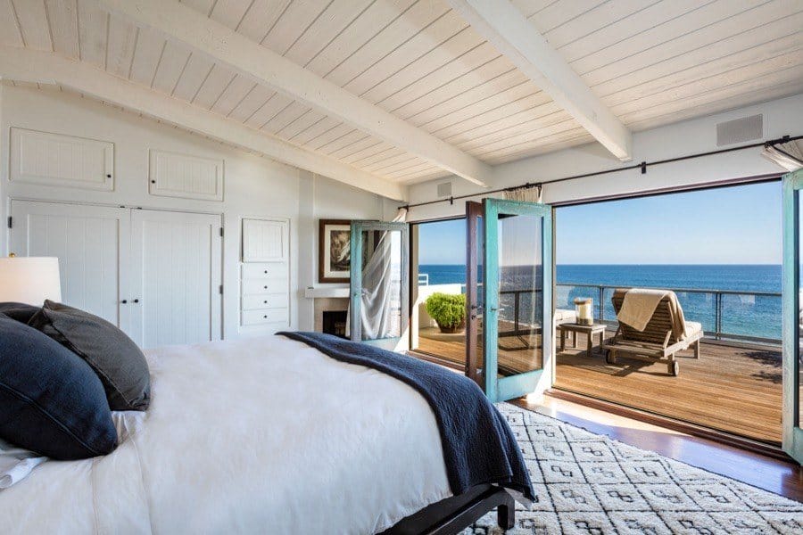 Bright bedroom with ocean view, open doors to a balcony, and cozy seating area on a sunny day.