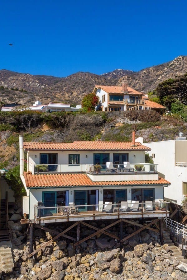 Oceanfront house on rocky shore with multiple balconies, blue windows, and mountain backdrop under a clear blue sky.