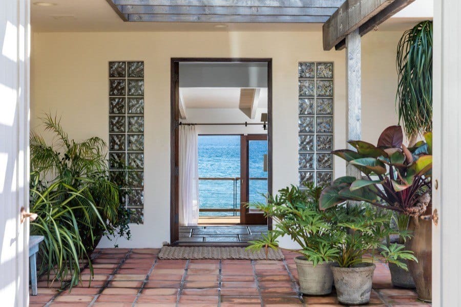 View through an open doorway leading to a balcony overlooking the ocean, surrounded by potted plants and natural light.