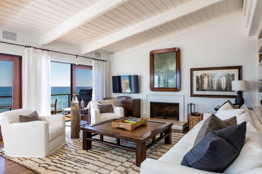 Cozy living room with ocean view featuring white sofas, wooden ceiling beams, and a fireplace.