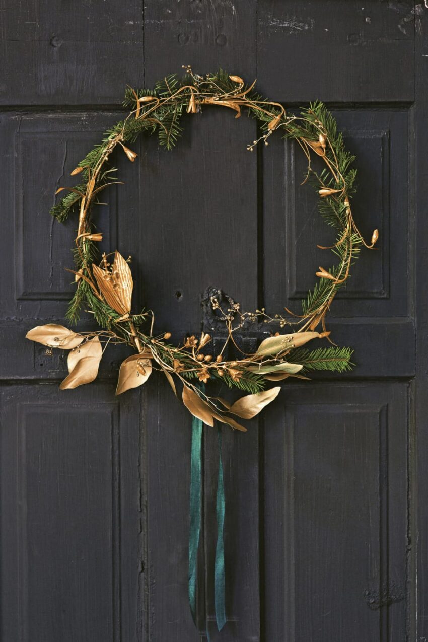 A minimalist wreath with greenery and gold decorations hanging on a dark wooden door.