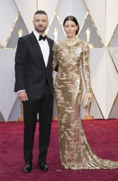 A couple on the red carpet, with the woman in a gold dress and the man in a black tuxedo at an awards event.