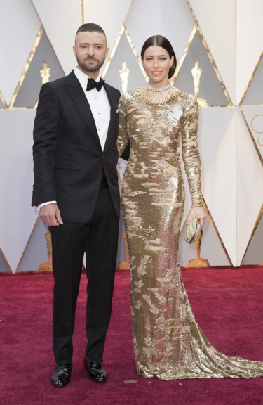 A couple on the red carpet, with the woman in a gold dress and the man in a black tuxedo at an awards event.
