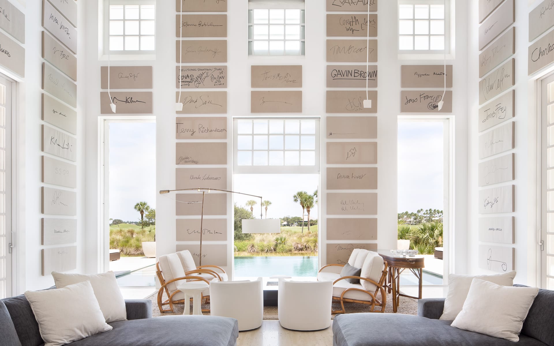 Spacious living room with large windows, signature-covered walls, gray sofas, white chairs, and a view of a pool and trees.