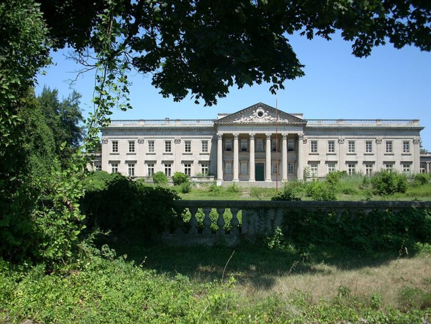 Grand historic mansion with tall columns and intricate architecture surrounded by lush greenery and a stone fence.