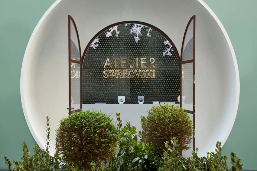 Elegant storefront entrance with Atelier Swarovski signage, framed by circular arch and lush greenery in foreground.