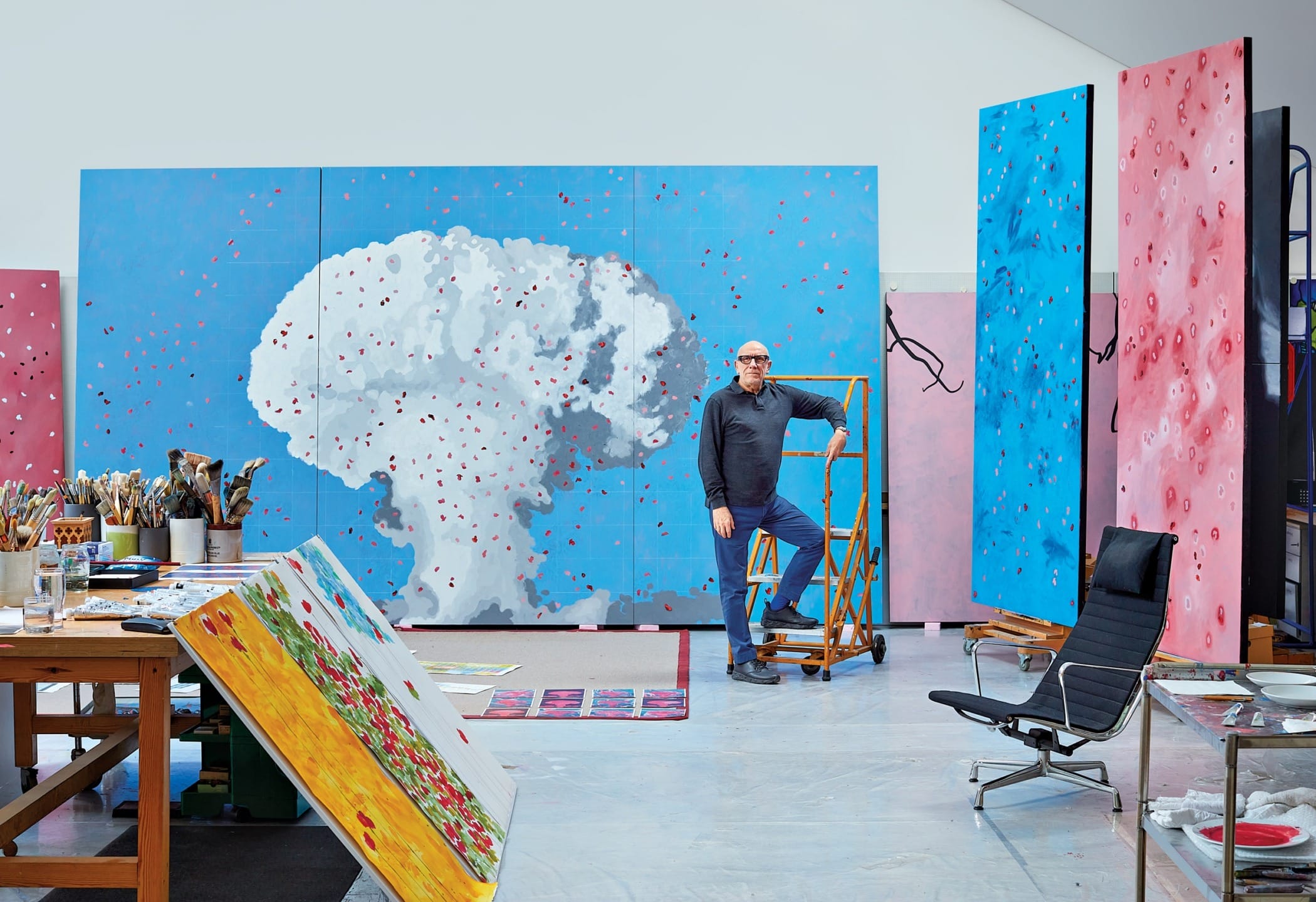 Artist standing in a colorful studio with large abstract paintings and art supplies around.