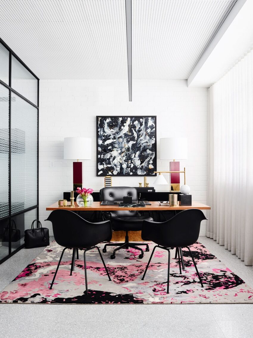 Modern office with abstract art, black chairs, wooden desk, decorative lamps, and a colorful rug in a bright, airy space.