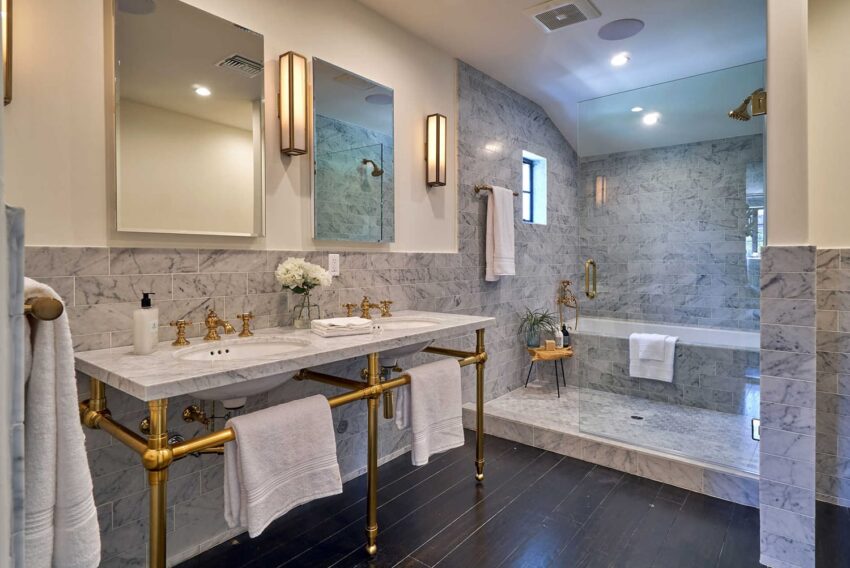Luxury bathroom with marble countertops, gold fixtures, glass shower, and wooden floor.