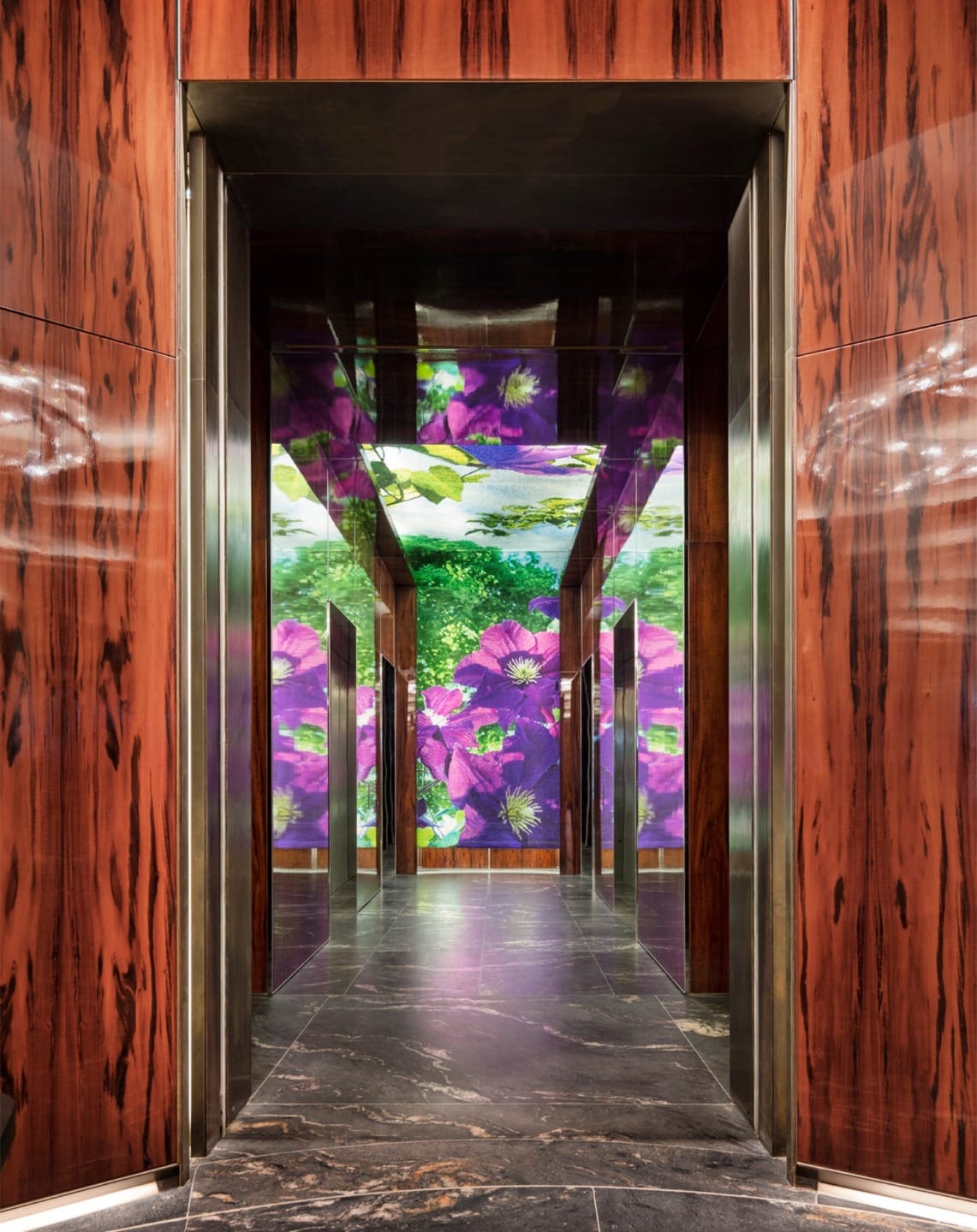 Elevator interior with floral digital art on panels and mirrored ceiling, surrounded by wood accents and dark marble floor.
